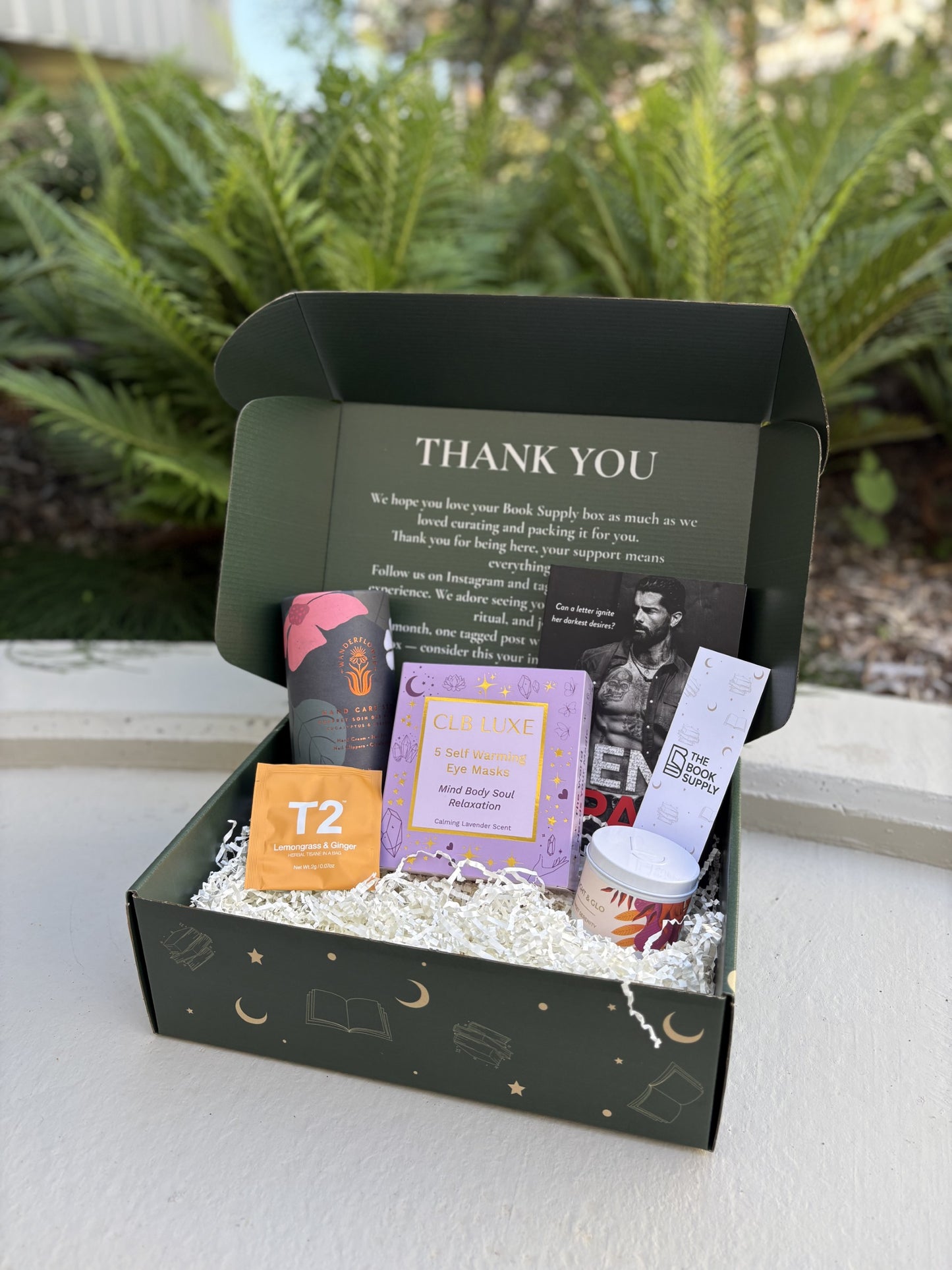Gift box with various items including a book and a bottle, set against a blurred green outdoor background.
